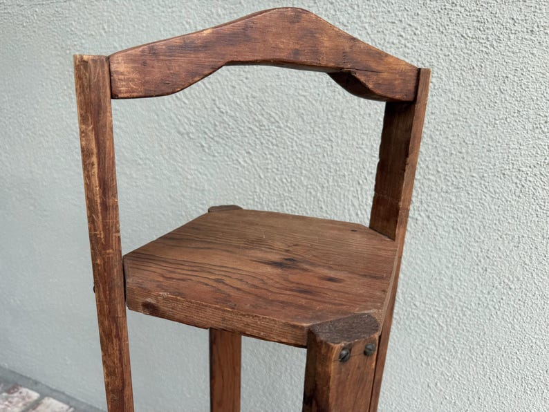 May include: A small, rustic wooden corner shelf with a triangular seat and a curved backrest. The shelf is made of dark brown wood and has visible wood grain. The construction features visible nails and a simple, functional design.