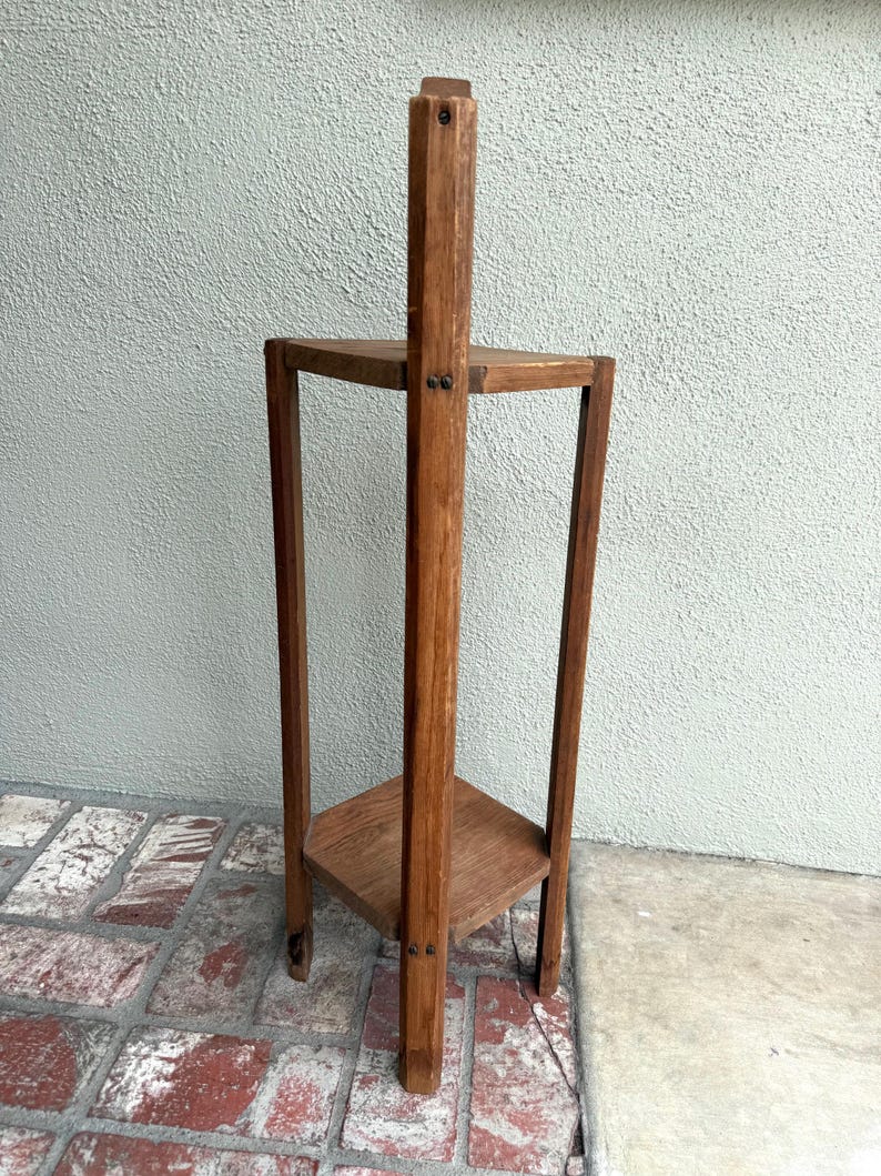 May include: A three-tiered, wooden plant stand with a natural finish. The stand has a central vertical support and three square shelves. The stand is approximately 76 cm tall and 30 cm wide.