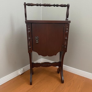 May include: A dark cherry-stained wooden cabinet with a handle, door, and decorative legs. The cabinet has a scalloped bottom edge and ornate detailing on the sides and door. The handle is a horizontal bar with carved details.