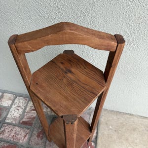 May include: A three-tiered, corner plant stand made of light brown wood. The stand has a triangular shape with three shelves and a decorative top. The wood grain is visible, giving it a rustic appearance. The stand is designed to fit into a corner.