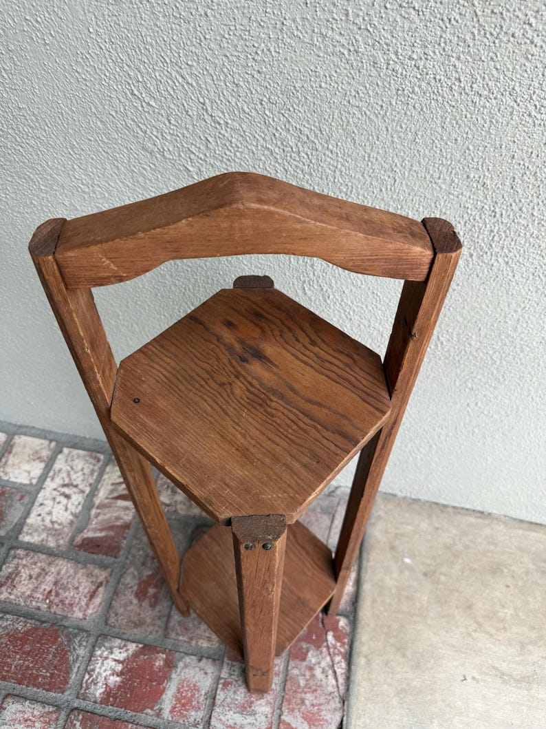 May include: A three-tiered, corner plant stand made of light brown wood. The stand has a triangular shape with three shelves and a decorative top. The wood grain is visible, giving it a rustic appearance. The stand is designed to fit into a corner.