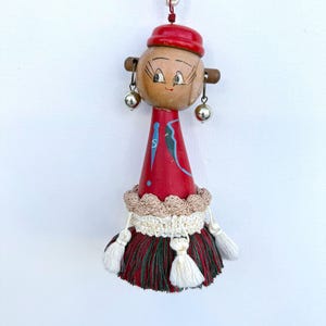 May include: A handmade wooden doll ornament with a red hat and dress. The doll has painted facial features, wooden earrings, and silver bell accents. The skirt is a mix of red, green, and white tassels, with a cream-colored trim. A white cord is attached for hanging.