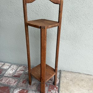 May include: A tall, wooden plant stand with three triangular shelves. The stand has a curved top and is made of a warm-toned wood. The shelves are supported by vertical posts, creating a simple, yet elegant design.