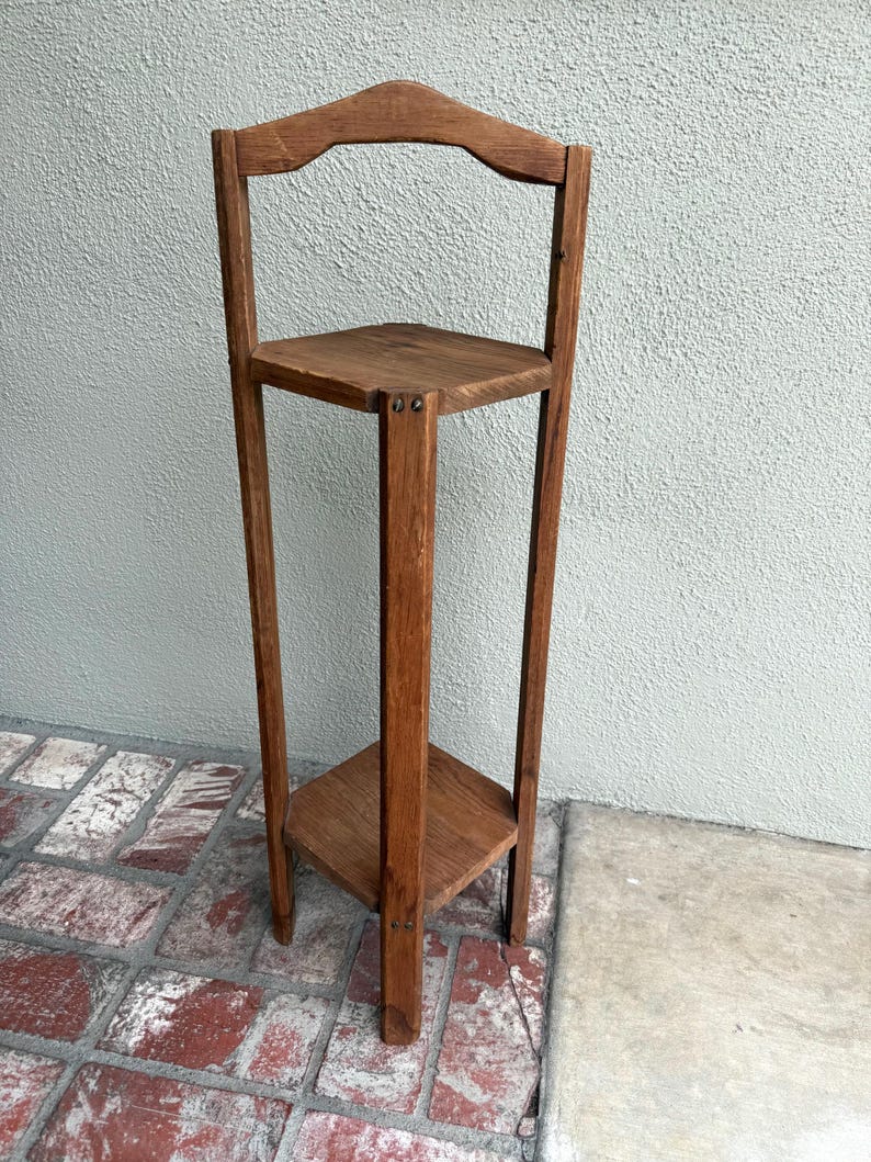 May include: A tall, wooden plant stand with three triangular shelves. The stand has a curved top and is made of a warm-toned wood. The shelves are supported by vertical posts, creating a simple, yet elegant design.