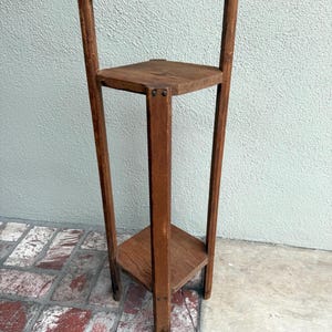 May include: A vintage wooden plant stand with a square top and bottom shelf. The stand has a tall, slender design with a decorative arched back. The wood has a warm, natural finish, showcasing the grain. It is a charming piece for displaying plants or decor.