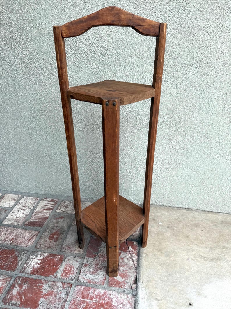 May include: A vintage wooden plant stand with a square top and bottom shelf. The stand has a tall, slender design with a decorative arched back. The wood has a warm, natural finish, showcasing the grain. It is a charming piece for displaying plants or decor.