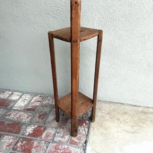 May include: A tall, vintage wooden plant stand with two triangular shelves and a central support beam. The stand is made of dark-stained wood and has a simple, geometric design. The stand is approximately 91 cm tall.