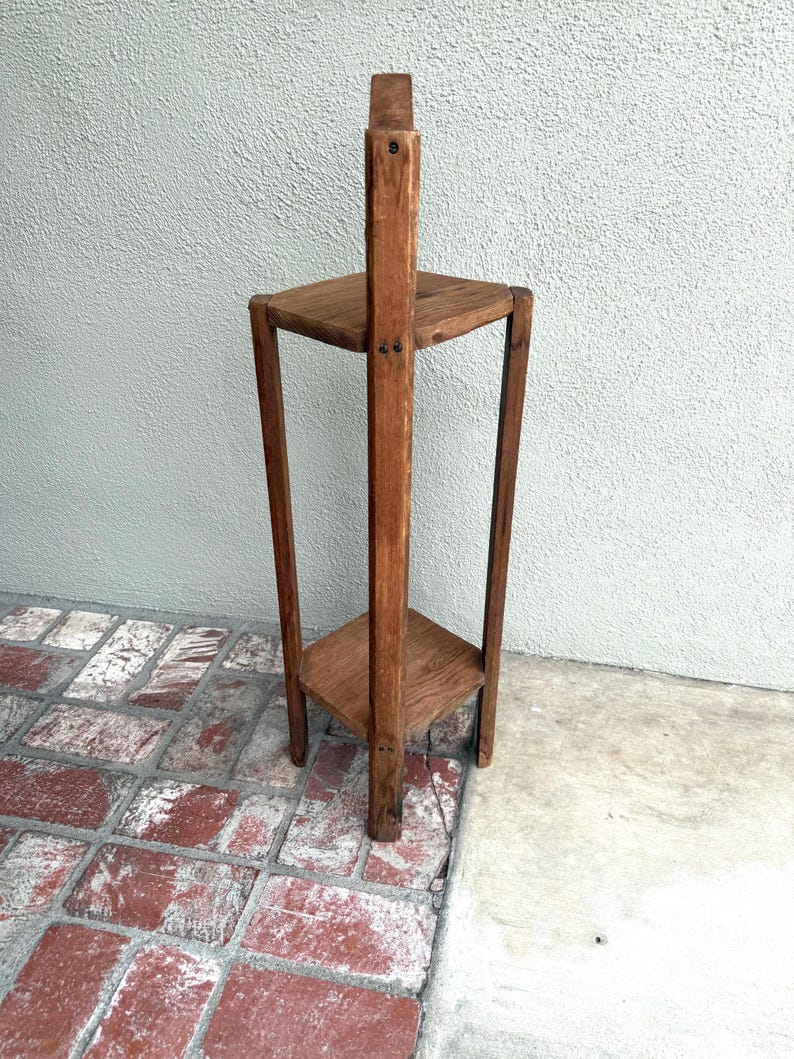 May include: A tall, vintage wooden plant stand with two triangular shelves and a central support beam. The stand is made of dark-stained wood and has a simple, geometric design. The stand is approximately 91 cm tall.
