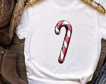 May include: White t-shirt featuring a hand-drawn candy cane design. The candy cane is red and white, with a curved shape. The shirt is displayed on a wooden surface with a gold hanger.