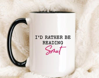 May include: White ceramic mug with a black handle and rim. The mug features the text "I'D RATHER BE READING Smut" in black and pink lettering. The mug is set against a soft, white, textured background.