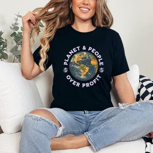 May include: A black t-shirt featuring a graphic of a globe and the text "Planet & People Over Profit". The person wearing the shirt is sitting on a white sofa.