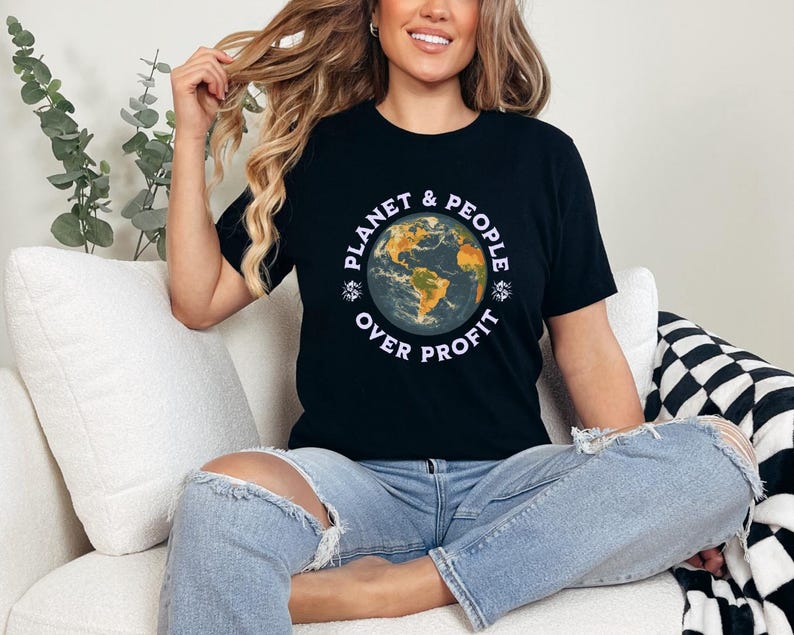 May include: A black t-shirt featuring a graphic of a globe and the text "Planet & People Over Profit". The person wearing the shirt is sitting on a white sofa.