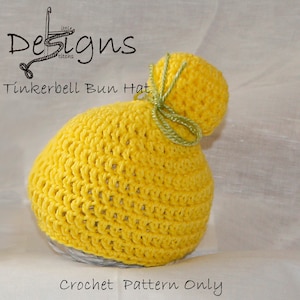 Puede incluir: Un gorro de ganchillo amarillo hecho a mano con una parte superior en forma de moño, atado con una cinta verde. El gorro está etiquetado como "Tinkerbell Bun Hat" y "Crochet Pattern Only". El diseño presenta un patrón de puntadas texturizadas.