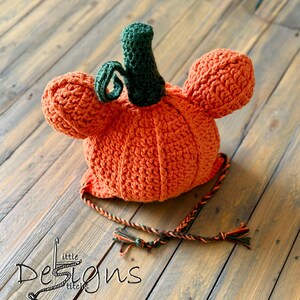 May include: An orange crochet pumpkin hat with a green stem and tendrils. The hat has ear flaps and a braided tie. The hat is handmade and perfect for fall or Halloween. The hat is sitting on a wooden surface.