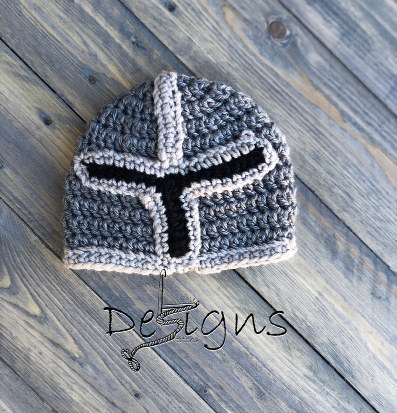 Puede incluir: Un gorro de punto de color gris y negro con forma de casco de caballero. El gorro tiene una visera negra y un patr&oacute;n gris y blanco.