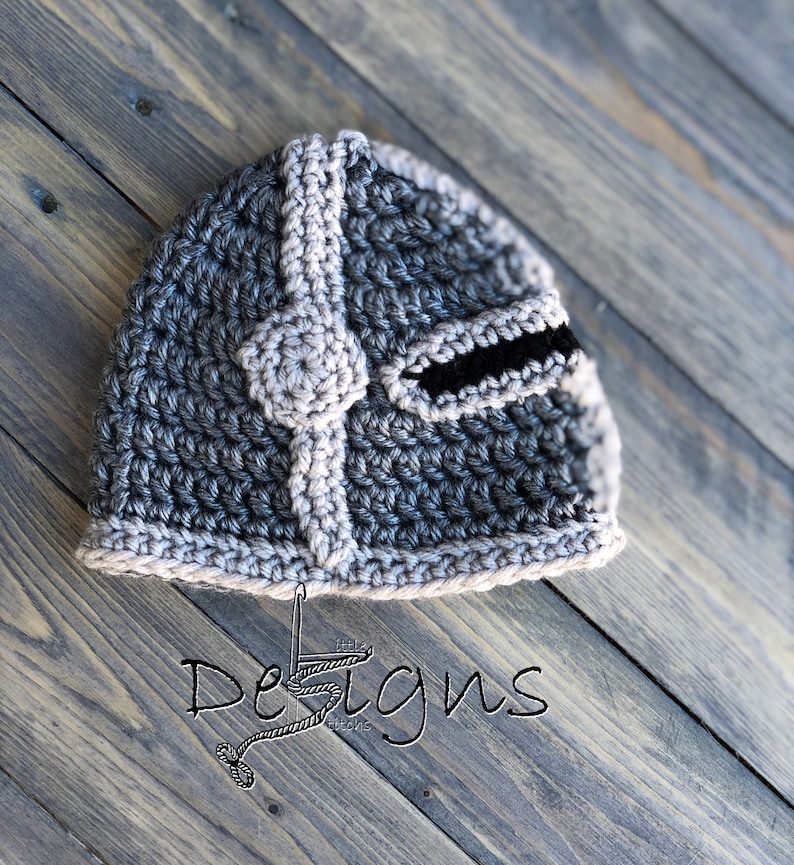 Puede incluir: Un casco de crochet gris y marr&oacute;n con una visera negra. El casco est&aacute; sobre una superficie de madera con el texto "Little Stitches Designs" en blanco.