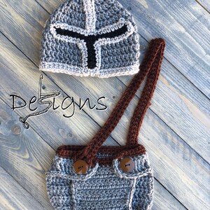Puede incluir: Sombrero de beb&eacute; de ganchillo gris con un dise&ntilde;o de casco de caballero blanco y negro. El sombrero se combina con un mono de ganchillo gris con tirantes marrones y botones marrones.