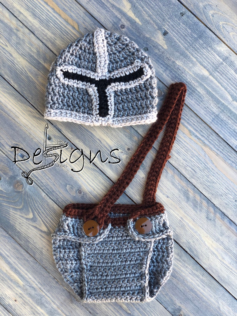 Puede incluir: Sombrero de beb&eacute; de ganchillo gris con un dise&ntilde;o de casco de caballero blanco y negro. El sombrero se combina con un mono de ganchillo gris con tirantes marrones y botones marrones.