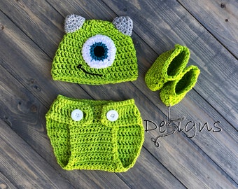 May include: A green crocheted baby outfit with a hat, diaper cover, and booties. The hat features a large white eye with a blue pupil and a green and white mouth. The outfit is inspired by the character Mike Wazowski from the movie Monsters, Inc.