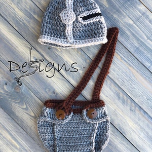 Puede incluir: Sombrero de beb&eacute; de ganchillo gris con dise&ntilde;o de casco y cubrepa&ntilde;al de ganchillo gris a juego con tirantes y botones marrones.
