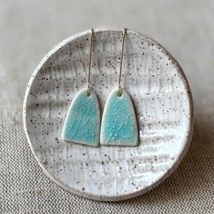 May include: A pair of artisan earrings with a light blue, crackled glaze. The earrings are arch-shaped and hang from gold-colored wires. They rest on a speckled, off-white ceramic plate, set against a neutral background.