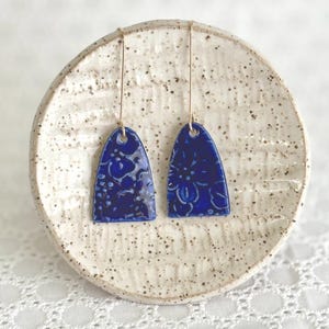 May include: A pair of cobalt blue ceramic earrings with a floral pattern. The earrings are teardrop-shaped with gold-colored ear wires. The earrings are displayed on a speckled, off-white ceramic plate.