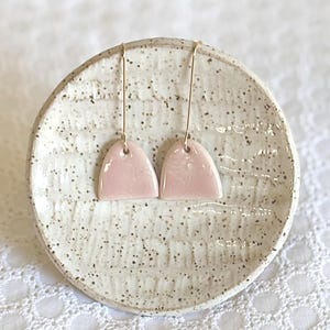 May include: A pair of pink ceramic earrings with gold hooks. The earrings are shaped like a half-circle and have a textured surface.