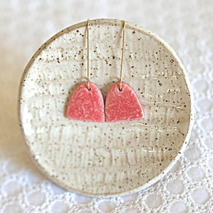 May include: A pair of pink ceramic earrings with gold hooks. The earrings are shaped like a rounded triangle and have a textured surface.