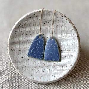 May include: A pair of blue ceramic earrings with a floral pattern, hanging from gold-colored wires. The earrings are displayed on a speckled white ceramic plate, set against a neutral background. The earrings have a unique, handcrafted appearance.