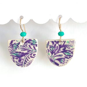 May include: A pair of ceramic earrings with a white base and a purple and green floral pattern. The earrings are shaped like a half-circle and have a gold hook with a turquoise bead.