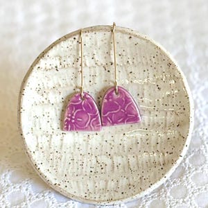 May include: A pair of purple ceramic earrings with a floral pattern. The earrings are hanging from gold hooks.