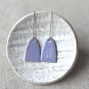May include: A pair of lavender ceramic earrings with a floral design, displayed on a speckled white ceramic plate. The earrings are a soft, muted purple color and hang from delicate gold-colored wires. The plate has a textured surface.