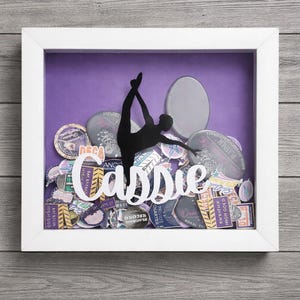 Puede incluir: Una caja de sombra con marco blanco, con un fondo púrpura y una silueta de gimnasta en split. El nombre "Cassie" está en escritura blanca. La caja está llena de varios parches y medallones coloridos.