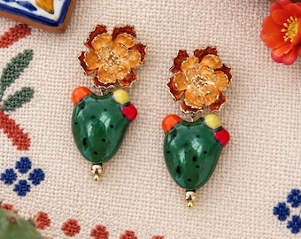 May include: A pair of dangle earrings featuring a gold-tone metal flower at the top and a green enamel cactus pendant below. The cactus has small black dots and colorful red and yellow accents. The earrings have a polished finish and gold hardware.