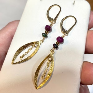 May include: Gold earrings with a leaf design and a dangling chain. The earrings are adorned with small red and green gemstones.