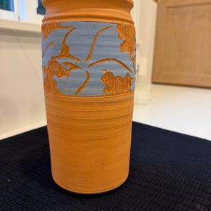 May include: A cylindrical terracotta vase with a matte finish and a flared rim. The upper section features a wide, horizontal band of light blue glaze with carved, leaf-like patterns that reveal the natural orange-brown clay underneath.
