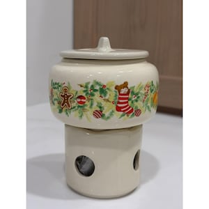May include: A cream-colored ceramic tea warmer with a removable lid. The body features a festive holiday print with a gingerbread man, a teddy bear in a red and white striped stocking, candy canes, ornaments, and green pine branches.