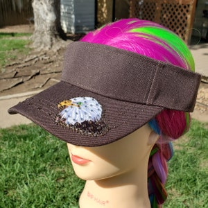 May include: A brown visor with a white and brown eagle design with rhinestones. The eagle has a green eye.
