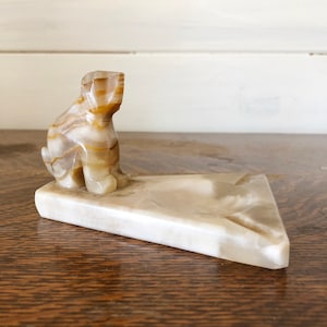 May include: A white marble ashtray with a carved dog figurine on the side. The ashtray is triangular in shape and has a small, round depression in the center.