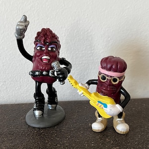 May include: Two vintage California Raisins figurines. One raisin figure is holding a microphone and has a raised arm. The other raisin figure is wearing glasses and a headband, holding a yellow guitar. Both figures are in a rock and roll pose.