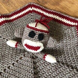 May include: A gray crocheted baby blanket with a red and white border. A crocheted monkey toy is attached to the blanket. The monkey has a red mouth, black eyes, and a white hat.