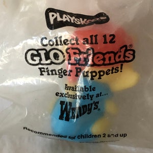 Op de afbeelding: Een plastic zak met een veelkleurige vingerpop. De pop is rood, geel en blauw. De zak heeft de tekst "PLAYSKOOL", "Collect all 12 GLO Friends Finger Puppets!", "Available exclusively at... WENDY'S", en "Recommended for children 2 and up".
