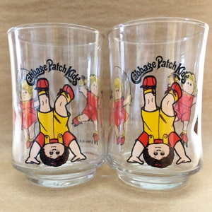 May include: Two clear glass Cabbage Patch Kids drinking glasses with a cartoon image of a Cabbage Patch Kid doing a handstand. The glasses have the words "Cabbage Patch Kids" printed on them.