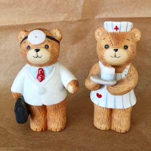 May include: Two ceramic teddy bear figurines, one dressed as a doctor with a stethoscope and a briefcase, the other dressed as a nurse with a red cross on her hat and a tray with a cup.