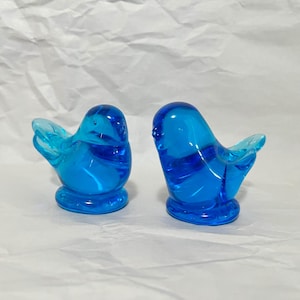 May include: Two blue glass bird figurines. Each bird has a rounded body, a small base, and outstretched wings. The glass has a swirl pattern, and the color is a vibrant shade of blue.