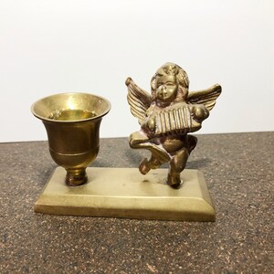 May include: A brass candle holder featuring a cherub playing an accordion. The holder has a single candle cup and a rectangular base.
