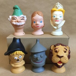May include: Six plastic character heads from the Wizard of Oz. The heads are of Dorothy, the Scarecrow, the Tin Man, the Cowardly Lion, the Wicked Witch of the West, and the Wizard. The heads are all in different colors and have different facial expressions.