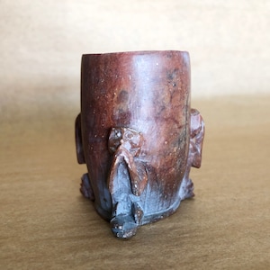 May include: A small, dark brown, wooden cup with a carved design of three figures. The cup is made of a dark wood with a smooth finish.