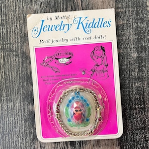 May include: Vintage Jewelry Kiddles bracelet by Mattel. The packaging is pink and white, with the text "Jewelry Kiddles" and "Real jewelry with real dolls!". The bracelet is silver-toned with a flower charm containing a small doll.