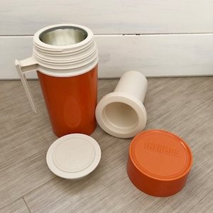 May include: An orange thermos with a white lid and a white inner lid. The thermos has a white handle and the word "THERMOS" is printed on the lid.